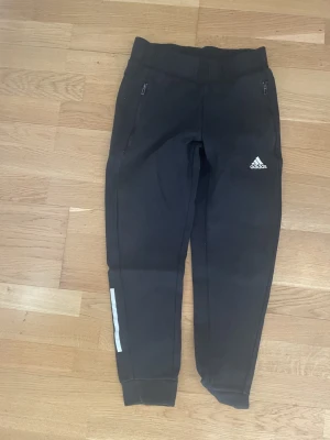 Svarta Adidas mjukisbyxor - Säljer ett par svarta mjukisbyxor från Adidas med vita detaljer och dragkedjefickor. Byxorna har en smal passform, mudd vid benslut och Adidas-logga på låret. Perfekta för chill eller träning.
