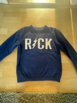 Blå stickad tröja ROCK Zadig & Voltaire - Mörkblå stickad tröja från Zadig & Voltaire med vit text 'ROCK' och blixt på bröstet. Tröjan har rund hals, långa ärmar och ribbstickade muddar. Perfekt för dig som gillar en cool och avslappnad stil.