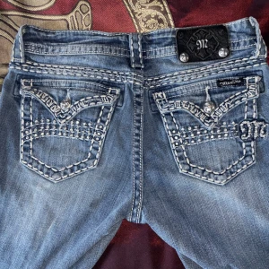 Miss Me jeans - Snygga blå jeans från Miss Me med kraftiga vita broderier och diamanter på bakfickorna. Jeansen har låg midja, klassisk femficksdesign och dekorativa detaljer längs sömmarna. Perfekta för dig som gillar statement-jeans med unik look.