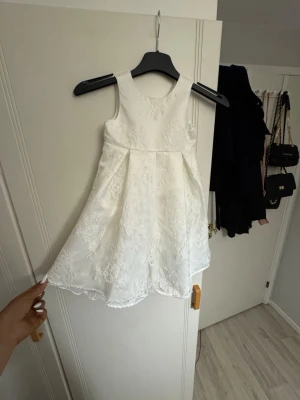 Vit ärmlös klänning med spetsdetaljer - Söt vit klänning med blommiga spetsdetaljer och ärmlös design. Klänningen har en lätt utsvängd kjol och rund halsringning, perfekt för en fräsch och stilren look. Passar dig som gillar romantiska och eleganta plagg.