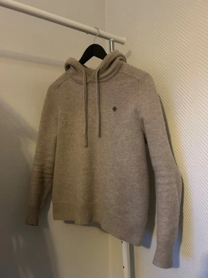 Beige stickad hoodie från Morris i merinoull  - Stickad beige hoodie från Morris med dragsko i huvan och diskret broderad logga på bröstet. Tröjan har långa ärmar och ribbade muddar vid ärmslut och nederkant. Perfekt för en avslappnad och stilren look. Sitter som S/XS