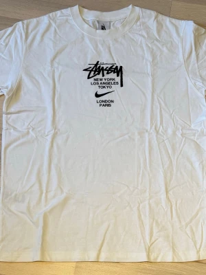 Vit Stüssy x Nike t-shirt - OBS, tröjan är storlek XL men passar som Medium, är 180cm och 65kg och den passar mig. Säljer en clean vit t-shirt från Nike i samarbete med Stüssy. Tryck på bröstet med Stüssy-logga och städer som New York, Los Angeles, Tokyo, London och Paris. Klassisk passform och rund hals. Perfekt till streetwear-stilen.