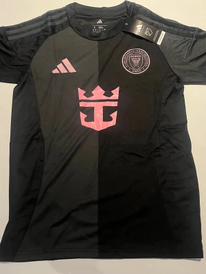 Svart Inter Miami tröja Adidas - Snygg svart kortärmad Inter Miami CF tröja från Adidas med rosa detaljer och klubbmärke på bröstet. Tillverkad i lätt och funktionellt material, perfekt för träning eller match. Klassisk Adidas-logga och rosa sponsortryck på framsidan.