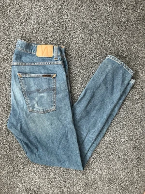 Blå jeans från Nudie - Säljer ett par klassiska blå jeans från Nudie med hög midja och raka ben. Jeansen har fem fickor, ljus tvätt och Nudie-logga på bakfickan. Perfekta för en avslappnad och stilren look.