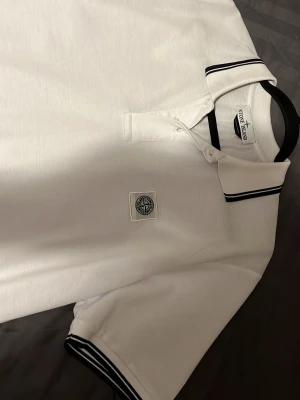 Vit pikétröja från Stone Island - Snygg vit pikétröja från Stone Island med svart-vita ränder på krage och ärmslut. Tröjan har klassisk pikékrage, två knappar och en broderad Stone Island-logga på bröstet. Tillverkad i mjuk bomull som känns skön mot huden.