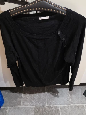 Svart stickad tröja från Stockh LM - Snygg svart stickad tröja från Stockh LM med rund halsringning och dekorativa knappar på axeln. Tröjan har långa ärmar och är tillverkad i ett mjukt, tunt material som passar perfekt till lager på lager-stil.