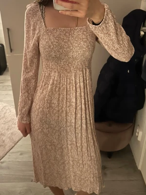 Ljusblommig långklänning med smock - Supersöt långklänning i ljusrosa med småblommigt mönster. Klänningen har smockad överdel, fyrkantig ringning och långa ärmar. Kjolen är lätt utställd och materialet känns mjukt och luftigt 