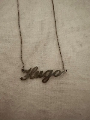 Silverfärgat namnhalsband Hugo - Snyggt halsband i silverfärg med namnet 'Hugo' i skrivstil. Kedjan är tunn och smidig, och namndetaljen är i metall med en blank finish. Perfekt accessoar för dig som vill ha ett personligt smycke med stilren design.