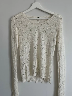 Vit stickad tröja med hålmönster - Snygg vit stickad tröja med lång ärm och dekorativt hålmönster i rutigt mönster. Tröjan har en rundad halsringning och är tillverkad i ett lätt och luftigt material, perfekt för lager-på-lager-stil.