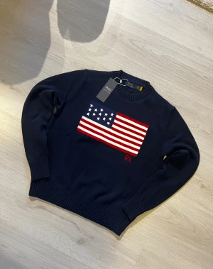  Polo Ralph Lauren tröja - O.B.S det står M på tagen men det passar i S. Snygg mörkblå stickad tröja från Polo Ralph Lauren med amerikansk flagga i rött, vitt och blått på bröstet samt RL-broderi. Tröjan har rund hals, långa ärmar och breda ribbade muddar vid ärmslut och nederkant. Perfekt för dig som gillar klassisk och cool stil.