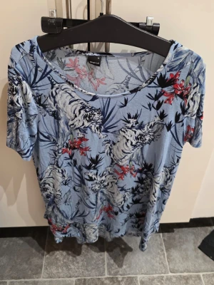 Blå topp  med tiger- och blommönster - Snygg blå topp från gina tricot med korta ärmar och coolt mönster av vita tigrar och röda blommor. Materialet känns mjukt och luftigt, perfekt för varmare dagar. Rund halsringning och normal passform.
