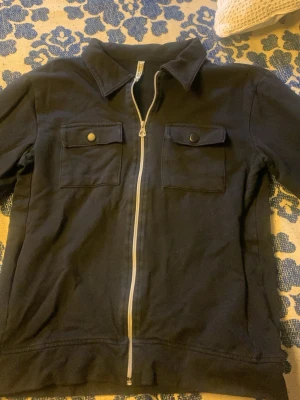 Svart overshirt med dragkedja - Stilren svart overshirt med hel dragkedja framtill och två bröstfickor med lock och knapp. Klassisk krage och långa ärmar med knapp vid ärmslut. Perfekt att slänga över en t-shirt för en avslappnad look.
