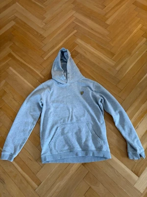 Ljusgrå hoodie från Lyle & Scott - Ljusgrå hoodie från Lyle & Scott med klassisk gul logga på bröstet. Hoodien har känguruficka framtill, ribbade muddar och en mjuk bomullskänsla. Perfekt för en avslappnad och stilren look.