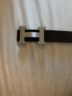 Svart skärp med H-spänne - Säljer ett klassiskt svart skärp från med det ikoniska H-spännet i silverfärgad metall. Bältet är stilrent och passar perfekt till jeans eller kostymbyxor. Materialet är skinn och spännet har en matt finish.