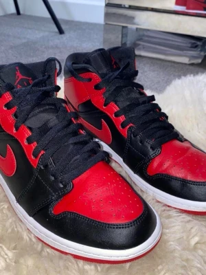 Air Jordan 1 Mid Svart/Röd - Säljer ett par Air Jordan 1 Mid sneakers i svart och rött läder med vita sulor och röda detaljer. Klassisk Nike Swoosh på sidan och Jumpman-logga på plösen. Skorna har svarta skosnören och perforerad tåbox för extra ventilation. ( UTAN BOXEN) 