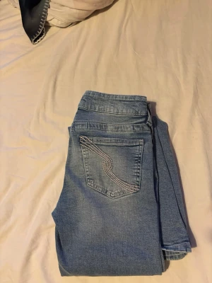 Ljusblå jeans från Gina Tricot - Säljer ett par ljusblå jeans från Gina Tricot med klassisk femficksdesign och snygg broderad detalj på bakfickan. Jeansen har bälteshällor och stängs med knapp och dragkedja. Perfekta för en avslappnad och trendig look.