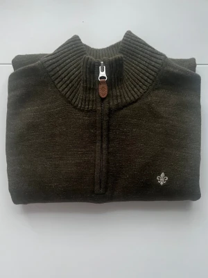 Olivgrön stickad tröja från Morris - Säljer en olivgrön stickad tröja från Morris med halv dragkedja och ribbad hög krage. Tröjan har lång ärm och en broderad vit lilja på bröstet. Perfekt för dig som gillar klassisk stil med en modern twist.inga hål