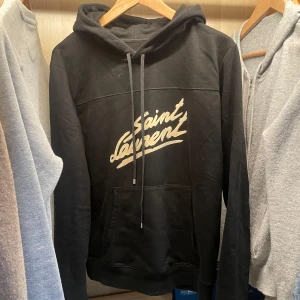 Svart hoodie från Saint Laurent - Snygg svart hoodie från Saint Laurent med deras ikoniska logga i vitt på bröstet. Tröjan är i toppskick bortsett från att den är lite smutsig på bilderna men tvättades strax efter så den är helt ren. Har haft denna i någon månad och säljer då den inte kommer till användning. Storlek S och är riktigt skön. Ej säker på äktighet, vad jag vet borde den vara 100% äkta men kan inte säkerställa. Tveka inte vid frågor
