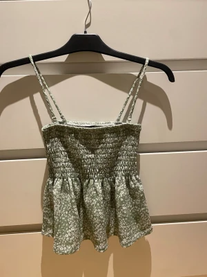 Grön blommig topp från Gina Tricot - Supersöt grön topp från Gina Tricot med små vita blommor. Toppen har smockad överdel, tunna axelband och volang nertill. 