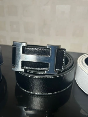 Svart skärp med H-spänne - Snyggt svart skärp med stort, silverfärgat H-spänne. Skärpet är gjort i skinn och har vita kontrastsömmar längs kanterna som ger en clean och exklusiv look. Perfekt accessoar för att lyfta vilken outfit som helst.