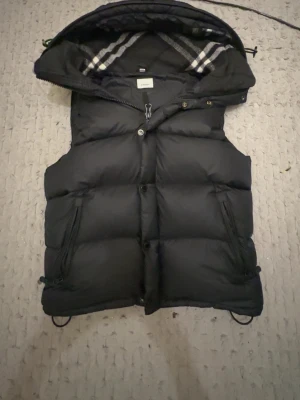 Svart dunväst med huva från Burberry - Snygg svart dunväst från Burberry med avtagbara ärmar och klassiskt rutigt foder i huvan. Västen har dragkedja och knappar framtill, två stora fickor med dragkedja och justerbar nederkant. Perfekt för lager-på-lager och streetstyle.