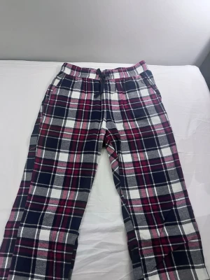 Rutiga pyjamasbyxor i flanell - Mysiga pyjamasbyxor i mjuk flanell med rutigt mönster i mörkblått, vinrött och vitt. Byxorna har resår i midjan och dragsko för extra komfort. Perfekta för chillkvällar hemma eller när du vill ha det extra bekvämt.