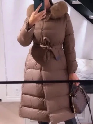 Brun lång dunjacka med pälskrage - Snygg brun lång dunjacka med markerad midja och knytskärp. Jackan har en stor pälskrage och quiltad design som ger extra värme. Perfekt för kalla vinterdagar och ger en trendig siluett.