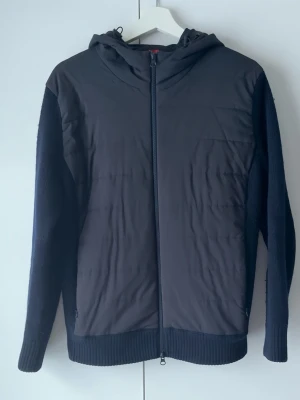 Jacka - Gran Sasso - Svart jacka med huva från Gran Sasso. kombinerar quiltad front i syntet med stickade ärmar och rygg. Dragkedja hela vägen och ribbade muddar. Perfekt för dig som gillar stilren och funktionell design.