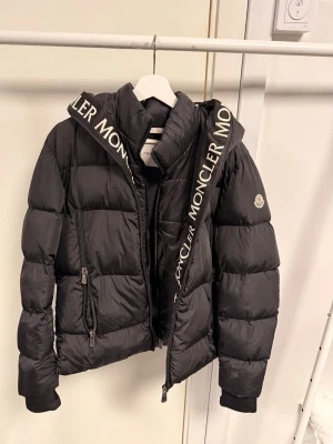 Svart pufferjacka från Moncler - Säljer en svart pufferjacka från Moncler med stor huva och tydlig logotyp på banden. Jackan har dragkedjor på fickorna och framtill, samt ribbade muddar vid ärmslut. Perfekt för kalla dagar och har en riktigt snygg streetstil.