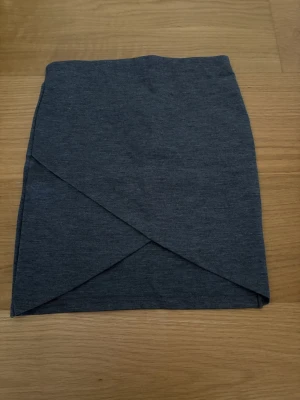 Mörkgrå omlottkjol från Gina Tricot - Säljer en mörkgrå kortkjol från Gina Tricot i omlottmodell. Kjolen har en stilren look med enkel design. Perfekt för dig som gillar minimalistisk stil och vill ha något som är lätt att matcha med olika toppar. 69% Polyester, 26% Viskos, 5% Elastan.