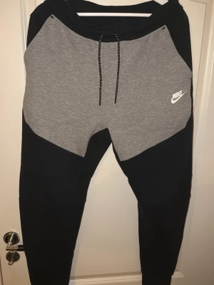 Nike tech äldre version mjukisbyxor svart och grå M - Nike tech mjukisbyxor i storlek M med svart och grå färgblock. Byxorna har snörning i midjan och en vit Nike-logga på framsidan. Materialet är mjukt och bekvämt, perfekt för chill eller träning. Snygg design med dragkedja på bakfickan för extra funktion. Knappt använda.