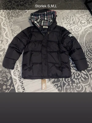 Svart Burberry pufferjacka med huva - Svart pufferjacka från Burberry med stor huva och klassiskt rutig insida i grått, vitt, svart och rött. Jackan har två framfickor med dragkedja och tydlig Burberry London England patch på ärmen. Perfekt för kalla dagar och riktigt snygg streetstil.