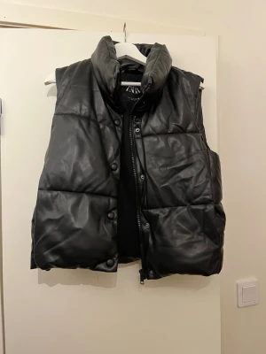 Svart skin väst från Zara - Säljer en svart skin väst från Zara i storlek S. Västen har hög krage, tryckknappar och dragkedja framtill. Materialet har en snygg skinnlook som ger en trendig vibe. Perfekt att slänga över en hoodie eller tröja!!🥰 västen är i ett välldigt bra skick!!