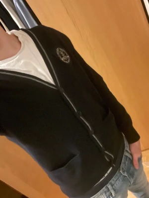 Mörkblå kofta - Snygg mörkblå kofta från LasseMaja med grå kantband och ett broderat emblem på bröstet. Koftan har v-ringning, knappar framtill och två fickor. Perfekt att slänga på över en t-shirt för en chill och stilren look.