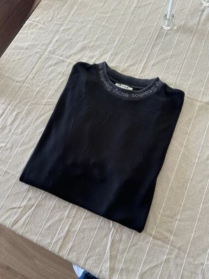 Svart t-shirt från Acne Studios - Svart t-shirt från Acne Studios med logotyp runt kragen. Modellen är enkel med rund hals och korta ärmar. Materialet är mjukt och stretchigt med en mix av viskos, nylon och elastan. Perfekt för dig som gillar stilrena och cleana plagg med diskret branding.