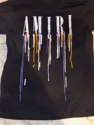 Svart AMIRI t-shirt med färgstänk - Svart t-shirt från AMIRI med stort tryck på ryggen och mindre tryck på bröstet. Trycket består av AMIRI-loggan med färgstänk i vitt, blått, rött och gult som rinner ner. T-shirten har rund hals och korta ärmar, tillverkad i mjuk bomull.