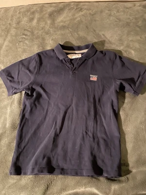 Mörkblå piké från Marqy Classic - Säljer en stilren mörkblå pikétröja från Marqy Classic med klassisk krage och korta ärmar. På bröstet finns en liten broderad amerikansk flagga och logotyp. Perfekt till jeans eller shorts för en avslappnad look.