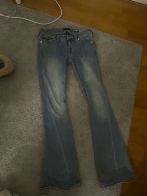 True religion Joey Low-Rise Flare Jeans - Säljer ett par ljusblå Joey low-rise flare jeans från true religon i storlek 26. snygga ficklock med knappar bak och slitningar framtill för en cool vintagekänsla. Perfekt för dig som gillar bootcut och låg midja.