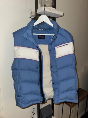 Blå dunväst Peak Performance  - Snygg blå dunväst från Peak Performance i modell Hudson V. Västen har vita paneler med röda detaljer upptill, hög krage och tryckknappar framtill. Tillverkad i polyamid med fyllning av dun och fjäder. Perfekt för lager-på-lager under kyliga dagar.