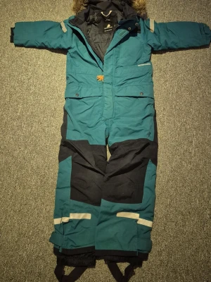 Turkos Didriksons vinteroverall 120 - Säljer en turkos och svart Didriksons vinteroverall med huva och fuskpäls. Overallen är 100% vattentät, har dragkedja, reflexdetaljer och justerbara benslut med resår, hela hällor. Lite skav finns längs ner på benslut men inget som påverkar funktionen. Lätt att laga.