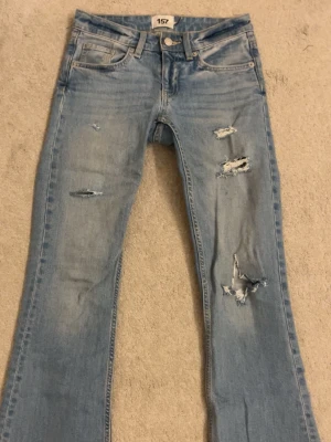 Bootcut ljusblå jeans med slitningar - Säljer ett par ljusblå jeans från 157 med bootcut-snitt och snygga slitningar på benen. Jeansen har klassisk femficksdesign, knapp och dragkedja i midjan. Materialet är jeans och passformen är normal med lite utsvängda ben. Midjemått är 34, är 158 cm