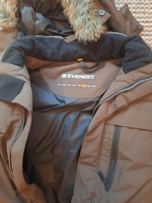 Brun parkajacka från Everest med fuskpäls - Brun parkajacka från Everest med huva och fuskpäls, flera praktiska fickor med dragkedja och stora framfickor. Jackan har lång ärm och är perfekt för kalla dagar. Materialet är polyester och syntet, och jackan har en klassisk parkamodell.