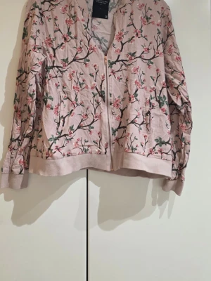 Blommig tröja från Lager 157 - Säljer en rosa tröja från Lager 157, The Black Label Collection. Tröjan har ett blommigt mönster med rosa och gröna detaljer, dragkedja framtill och ribbade muddar. Materialet är 100% viskos och modellen är lite loose fit. Perfekt för dig som gillar unik stil.