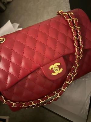 Röd quiltad axelväska  - Snygg röd axelväska  i quiltat skinn med gulddetaljer. Väskan har en klassisk design med en ikonisk logotyp på framsidan och en praktisk dragkedja inuti. Perfekt för att ge en touch av lyx till din outfit. Pris kan diskuteras vid snabbt köp!!