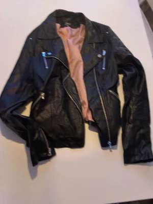 Svart skinnjacka med dragkedjor - Säljer en svart skinnjacka med coola silverfärgade dragkedjor och klassisk bikerkrage. Insidan har ett ljusrosa foder som ger en snygg kontrast. Perfekt för dig som vill ha en edgy look till vardagsoutfiten.