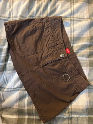 Brun kortkjol från Esprit med detaljer - Säljer en brun kortkjol från Esprit med coola fickor och dragkedjor både fram och bak. Kjolen har en snygg knapp med blomdetalj och en metallring på sidan. Tillverkad i bomull, perfekt för dig som gillar praktiska detaljer och en avslappnad stil. 40 cm rakt över 23 cm lång fram 27 cm lång bak. 