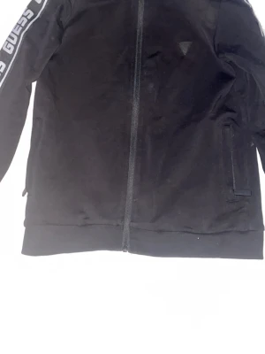 Svart zip hoodie från Guess - Svart hoodie från Guess med dragkedja framtill och logga på bröstet. Snygga vita GUESS-detaljer längs ärmarna. Tillverkad i mjuk bomullsmix och har långa ärmar samt två fickor på sidorna. Perfekt för en chill och sportig look.