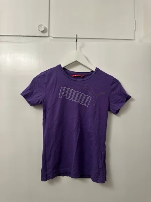 Lila Puma t-shirt i bomull - Snygg lila t-shirt från Puma med stort tryck på bröstet och klassisk logga både fram och bak. Skön passform och mjukt material som funkar perfekt till en avslappnad stil. Rund halsringning och korta ärmar.