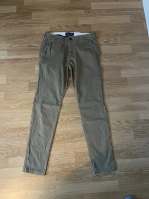 Beiga chinos från Abercrombie & Fitch - Säljer ett par beiga chinos från Abercrombie & Fitch i modellen Super Skinny. Byxorna har klassiska sidofickor, två bakfickor med knapp och diskret A&F-logga. Tillverkade i mjuk bomull med smal passform och midwaist. Perfekta för en clean och stilren look.