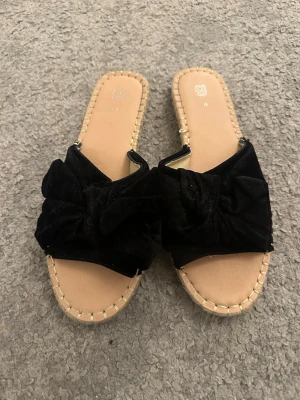 Svarta sandaler med rosett från Gina Tricot - Säljer ett par snygga svarta sandaler från Gina Tricot i storlek 38. De har en bred rosett framtill i mjuk sammet och en beige sula med dekorativt grovstickad kant. Platt sula och öppen tå gör dem perfekta för varma dagar.